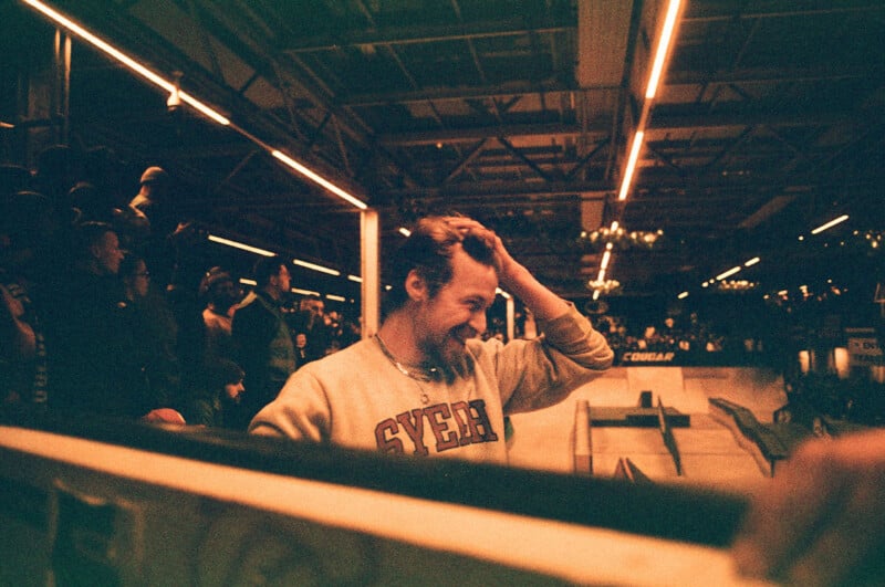 A person in a light sweater with "SYERA" on it stands in an indoor skatepark, smiling and brushing their hair back. The scene is warmly lit, with spectators in the background and a ramp visible on the right.