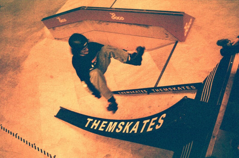 A person skateboarding in an indoor skatepark, performing a trick mid-air near ramps labeled "THEMSKATES." The scene is captured in a warm, vintage tone, emphasizing movement and action.