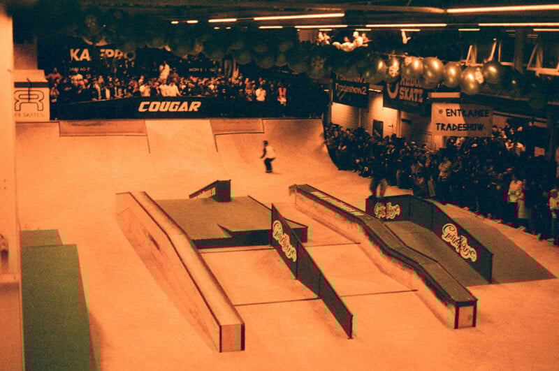 A vintage-style photo of an indoor skatepark competition. A skater performs on ramps and rails while a large crowd watches. The venue is decorated with banners and balloons, and the image has a reddish-brown tone.