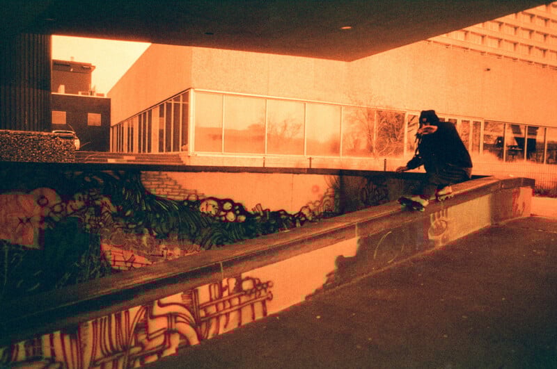 A person in a hat sits on the edge of a concrete structure covered with vibrant graffiti. The scene is bathed in an orange hue, giving it a moody, vintage feel. A large building with glass windows is in the background.