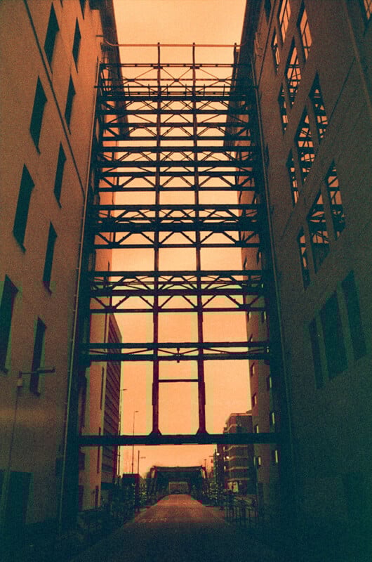 A narrow alley between two tall buildings with a series of metal beams crisscrossing overhead. The sky displays an orange hue, casting a warm glow on the scene. The path is empty, leading to an open area at the end.