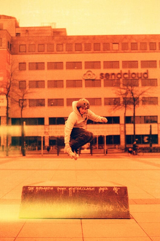 A person wearing a helmet and hoodie performs a jump on a skateboard over a concrete block in an urban area. The background features a building with the "Sendcloud" logo. The image has a retro, sepia-like color tone.