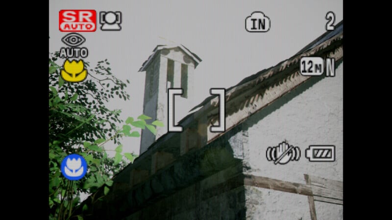 Camera viewfinder interface displaying a rustic building with a slanted roof and chimney. Lush green leaves are visible on the left. Various camera icons and settings are shown on the screen, including focus brackets and battery level.