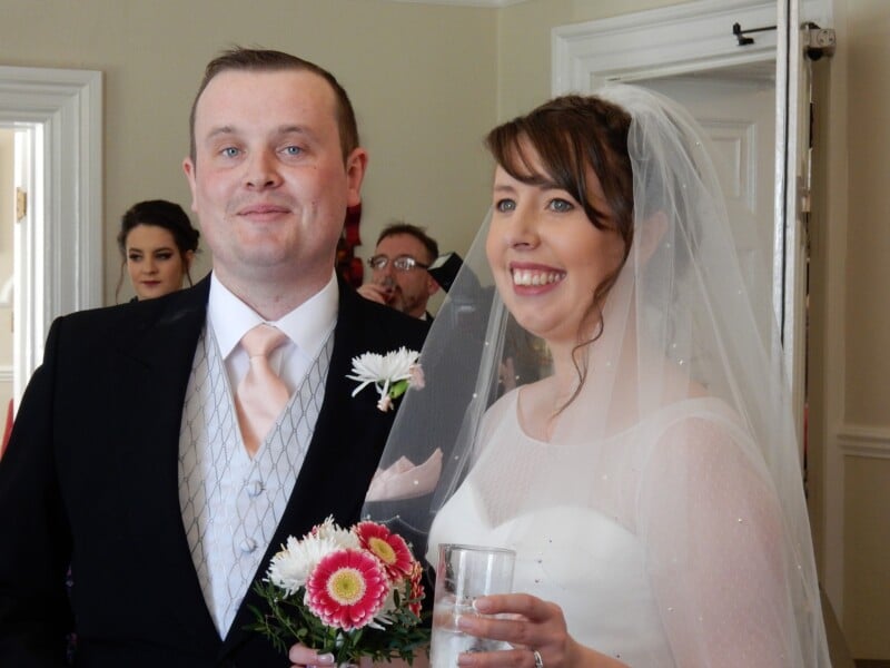 A smiling bride in a white dress and veil stands next to a groom in a suit holding a bouquet of pink and white flowers. Both are indoors, with a woman and other guests in the background.