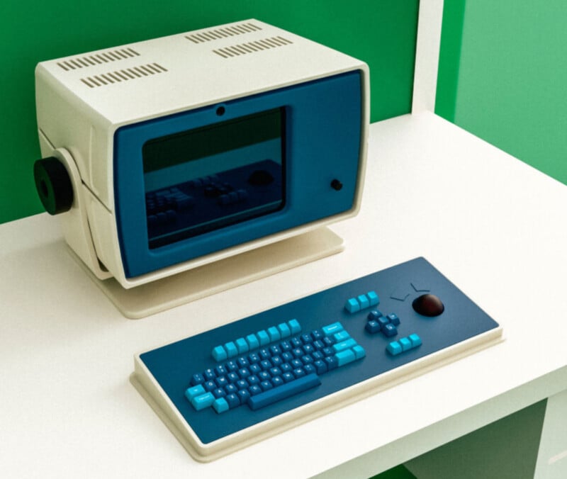 A retro-style computer with a cream and blue monitor and matching keyboard sits on a white desk against a green wall. The computer has a vintage design with vents and a knob on the side.