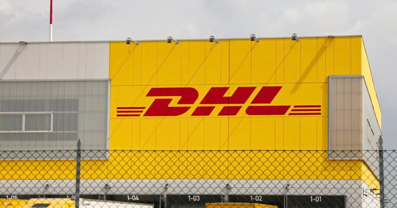 A large yellow DHL building with the company’s red logo on the side, viewed behind a chain-link fence under a cloudy sky.
