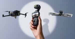 A hand holds a digital camera with a small screen, flanked by two flying drones against a neutral background with a circular spotlight.