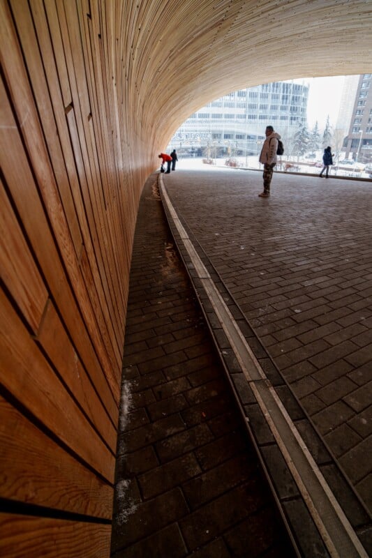 Wide-angle view of a tunnel with wooden walls and a brick path. A few people are walking and standing in the distance. The tunnel opens up to a cityscape with buildings visible under a cloudy sky.