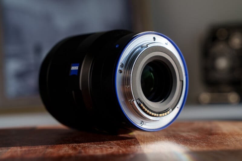 A close-up of a camera lens laying horizontally on a wooden surface. The metal lens mount is visible, with electronic contacts around the edge. The background is blurred, and sunlight is reflecting faintly off the surface.