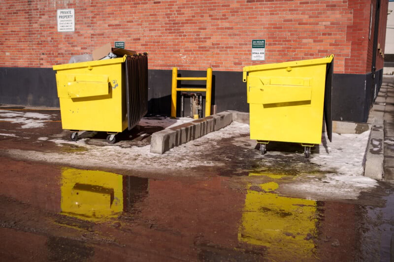 Two yellow dumpsters stand on a slightly wet, snowy area against a red brick wall. Reflections of the dumpsters are visible in the water on the ground. Signs labeled "Private Property" and caution signs are on the wall.