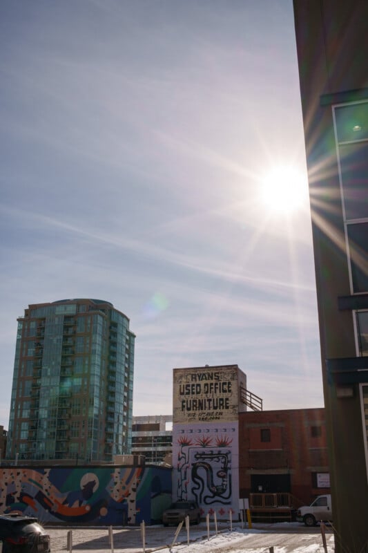 Sunlit urban scene with a tall residential building and a wall mural. The sign "Ryan's Used Office Furniture" is visible on a smaller building. The sky is clear, and the sun is shining brightly, casting long shadows on the almost empty street below.