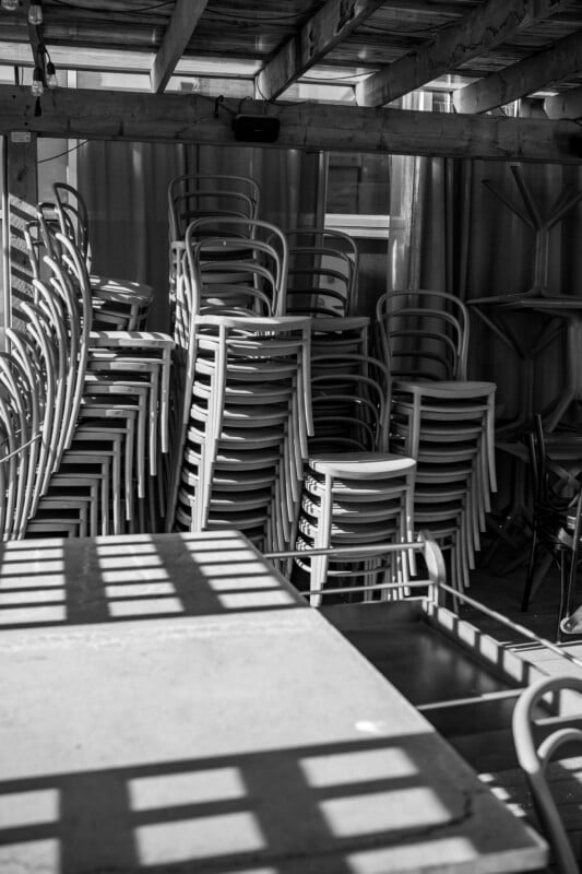 A black-and-white photo of several stacks of wooden chairs in a covered outdoor area. The sunlight casts a grid-like shadow on the floor and tables in the foreground, highlighting the patterns of light and shade.