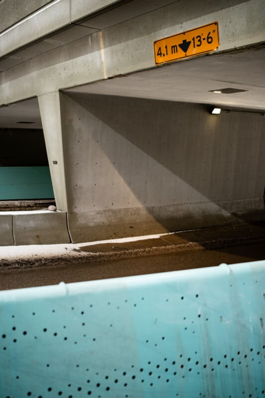 A sloped entrance to a parking garage with a clearance sign reading "4.1 m 13' 6". The area is surrounded by teal barriers, and the ground is wet with scattered snow. Overhead lights are illuminated, casting shadows along the walls.
