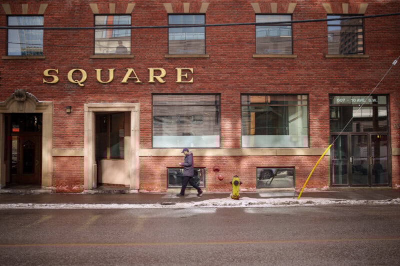 A person wearing a coat and hat walks past a red brick building with the word "SQUARE" in large gold letters. The street is wet and lightly dusted with snow. A fire hydrant and a yellow pole are visible along the sidewalk.