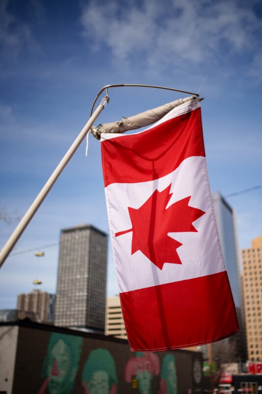 A Canadian flag with a red maple leaf flutters on a pole against a clear blue sky. In the background, several skyscrapers and buildings are visible, along with a mural on the side of a nearby structure.