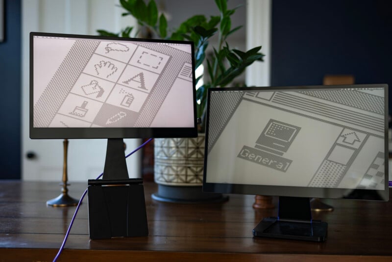 Two e-paper displays on a wooden desk show grayscale graphics and icons with plants in the background. One screen prominently displays geometric shapes and icons, while the other shows partial text and images. The scene is in a modern room.