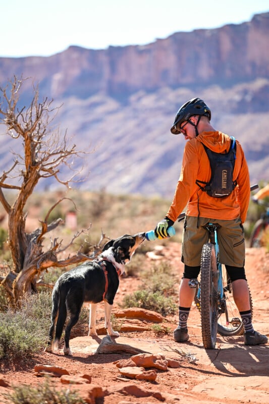 A cyclist in an orange jacket offers a water bottle to a black and white dog on a rocky trail. The cyclist stands beside a mountain bike, surrounded by a desert landscape with red cliffs and sparse vegetation.