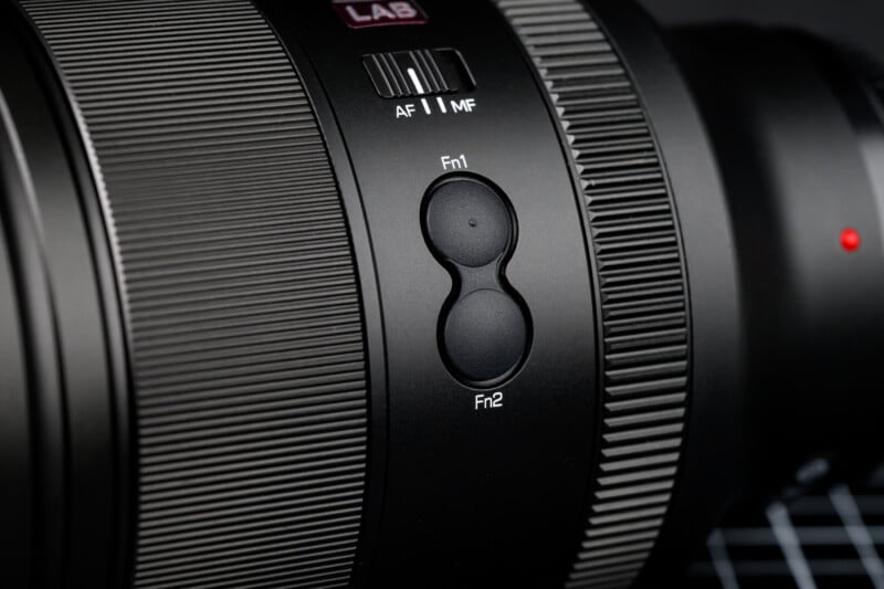 Close-up of a camera lens with visible focus modes labeled "AF" and "MF." Two round buttons labeled "Fn1" and "Fn2" are prominently displayed. The lens surface features textured grip rings and a small red dot for alignment.