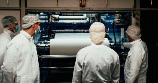 Several people wearing white lab coats, hairnets, and masks are observing a large industrial machine with rollers and white material. The setting appears to be a manufacturing facility or laboratory.