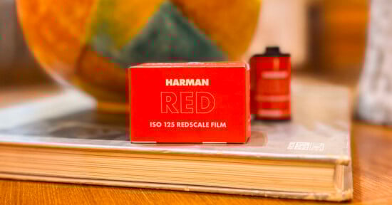 A red box of Harman Red ISO 125 Redscale film sits on a closed book with a blurred background. Next to it is a film roll, also blurred.