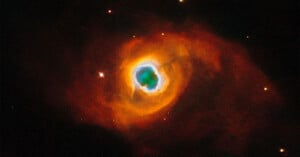 Bright nebula in space, showing vivid orange and red clouds surrounding a luminous blue and white center. Small distant stars are scattered across the dark background.