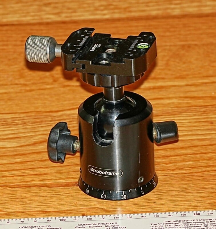 A black Stroboframe tripod head with adjustment knobs and a quick-release plate sits on a wooden surface. A ruler with metric and imperial measurements is partially visible at the bottom.