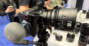 A close-up of a professional camera mounted on a tripod, equipped with a 35mm lens. In the background, people are holding phones and cameras. Various camera lenses are displayed on a table.
