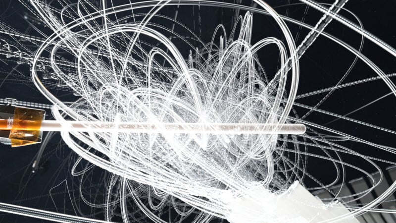 A long-exposure photo showing chaotic, swirling white light trails against a dark background, intersected by a horizontal rod with a metallic attachment on the left side.