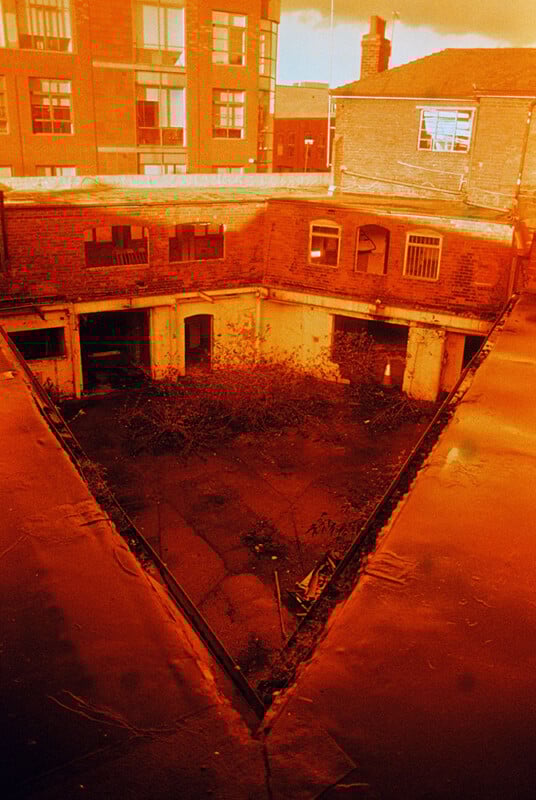 A vivid red, triangular-shaped courtyard surrounded by old brick buildings with windows. The courtyard is overgrown with weeds. The sky is a deep orange, casting a warm hue over the scene.