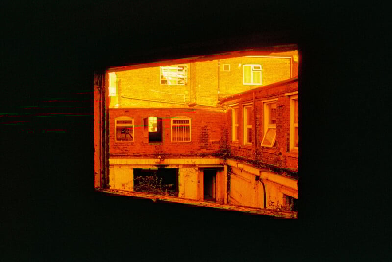 View through a dark, rectangular window of a brick building courtyard bathed in intense orange light. The scene includes windows with uneven frames and a partially overgrown ground. The light creates a warm, surreal atmosphere.