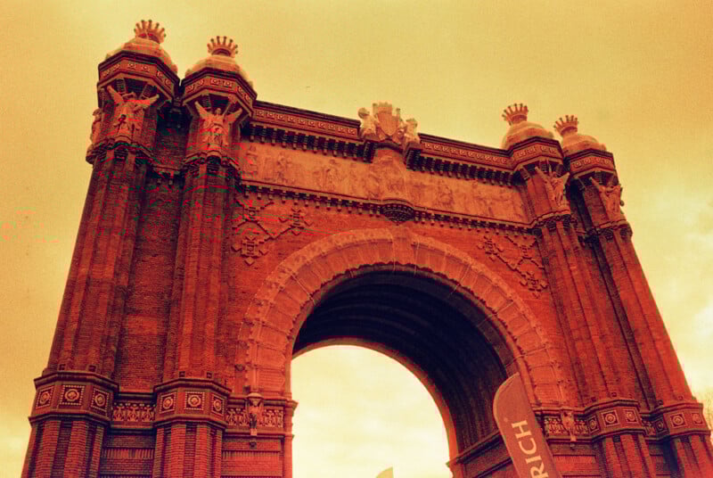 A sepia-toned image of a large, ornate archway with detailed stone carvings and decorative pillars. The sky is overcast, and there are partial banners visible at the bottom.