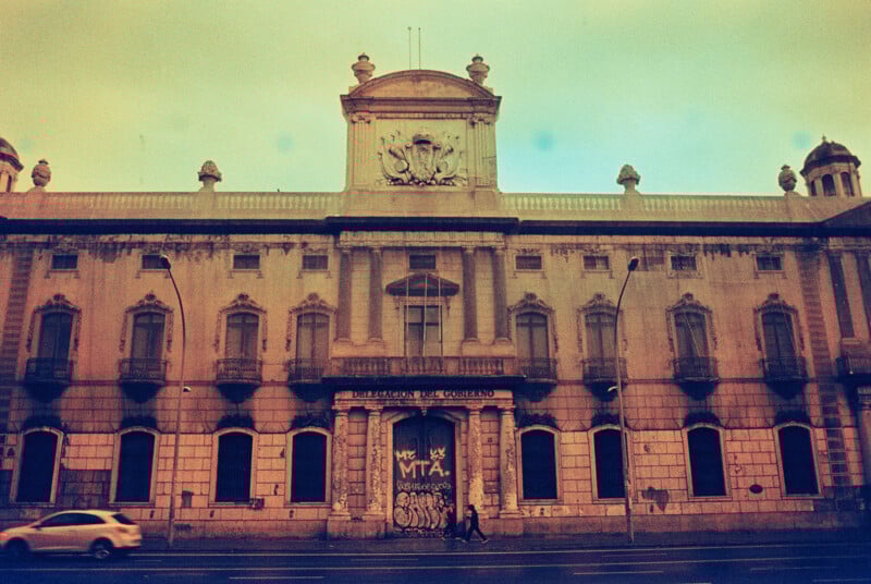 A vintage-style photo of an old, ornate building with arched windows and detailed stonework. The entrance is boarded up with graffiti. A person walks by on the street in front, and the building is slightly worn with age.