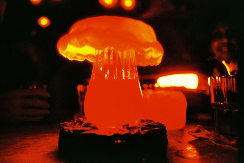 A glowing cocktail shaped like a mushroom cloud with a vibrant orange and red hue, set on a dark table. Other drinks and dim lights are visible in the out-of-focus background, creating an ambient atmosphere.