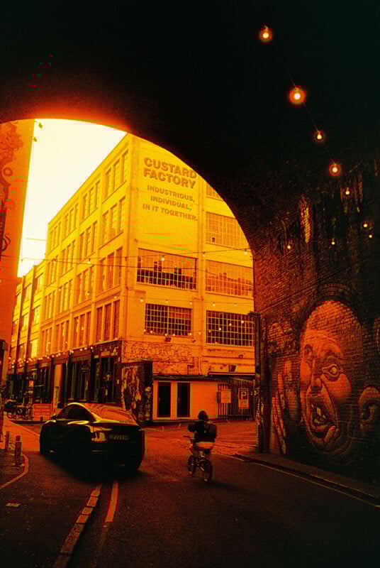 A vibrant orange-tinted photo shows a cyclist and car passing under a brick archway. To the right, a wall features a large mural of a face. In the background, there's a building with signs promoting industry and community.
