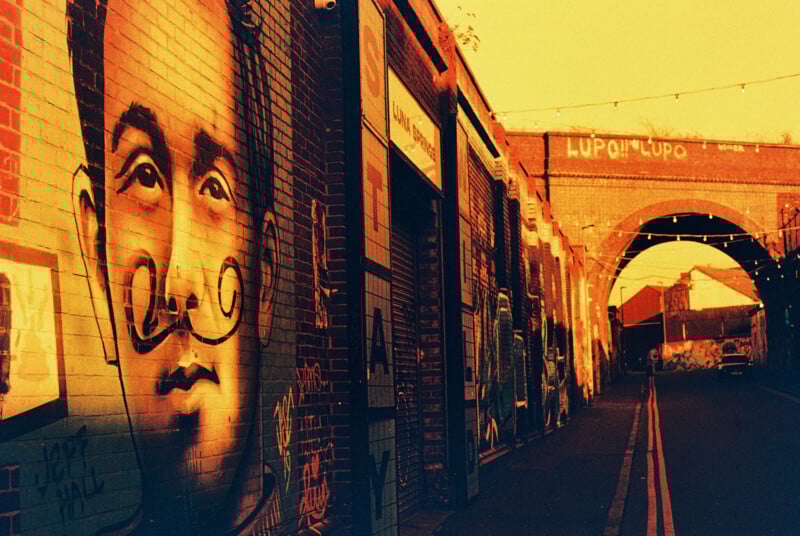 Street art mural of a man with a mustache on a brick wall at sunset. The image is in a warm, yellow tone. The street has string lights and leads to a brick archway in the background.