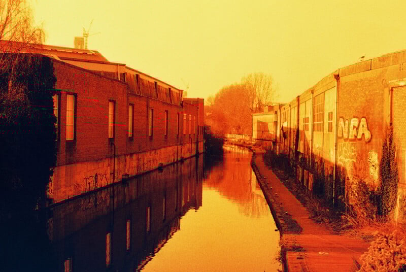 A canal lined with industrial buildings reflects a warm orange hue. Graffiti is visible on one of the buildings, and bare trees are in the background, capturing a serene and vintage urban scene.