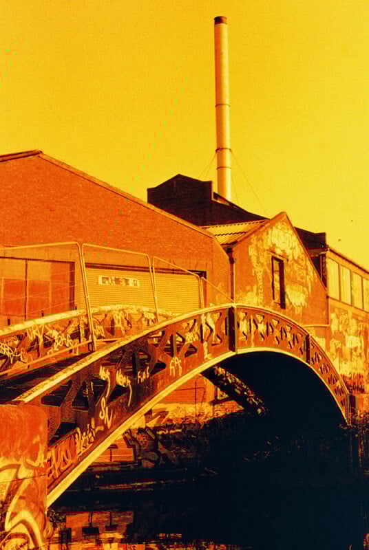 A vivid orange-tinted image shows a graffiti-covered bridge over a canal. In the background, an industrial-style building with a tall chimney is visible. The urban scene is bathed in warm light, emphasizing the vibrant street art.