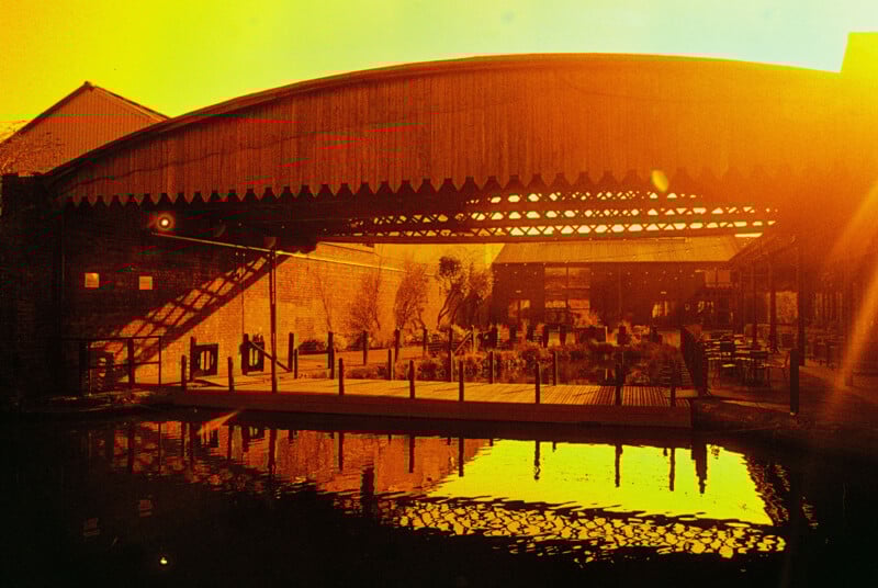 Sunlit industrial building with large overhang and seating area by a reflective water surface. Warm golden tones and architectural details create a nostalgic ambiance.