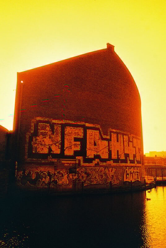 A large brick building with bold graffiti art covering its side is reflected in a body of water. The scene is bathed in an intense orange-yellow light, creating a dramatic, atmospheric effect.