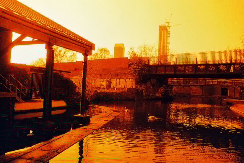 A canal with reflective water and graffiti-covered walls under a bright orange sky. A wooden structure is on the left, and a bridge is in the background with a construction crane visible. Sparse trees add texture to the urban scene.