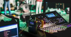 A sound mixing console with colorful buttons and displays is in focus. Nearby, a Saramonic audio receiver with antennas is attached. In the background, a blurred band setup is visible, indicating a live performance setting.