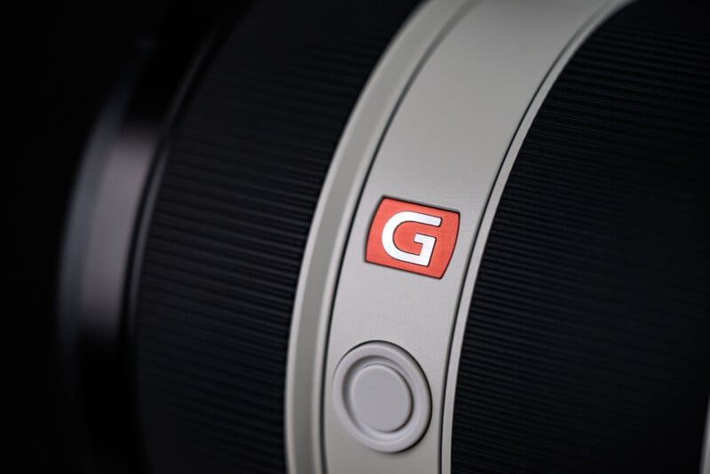 Close-up of a camera lens featuring a red "G" badge on a light gray ring, with a circular button below, and textured black focus and zoom rings on either side.