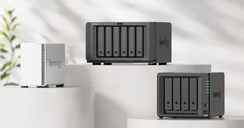 Three Synology NAS devices of varying sizes are displayed on white cylindrical pedestals against a light, blurred background with a hint of greenery. The devices feature multiple drive bays and indicator lights.