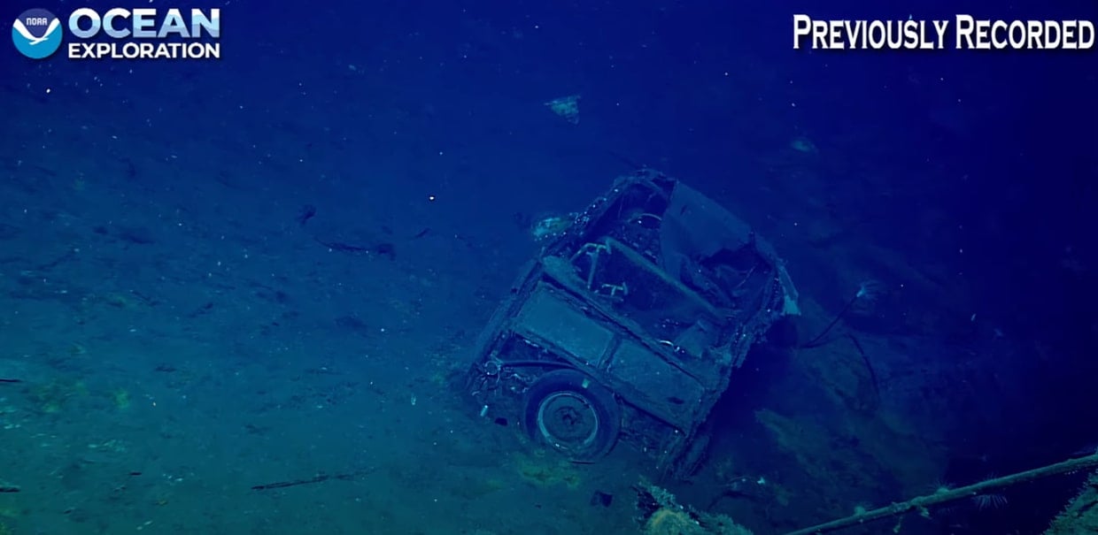 Underwater photo of a rusted, partially buried vehicle on the ocean floor, with the NOAA Ocean Exploration logo and “Previously Recorded” text in the corners.