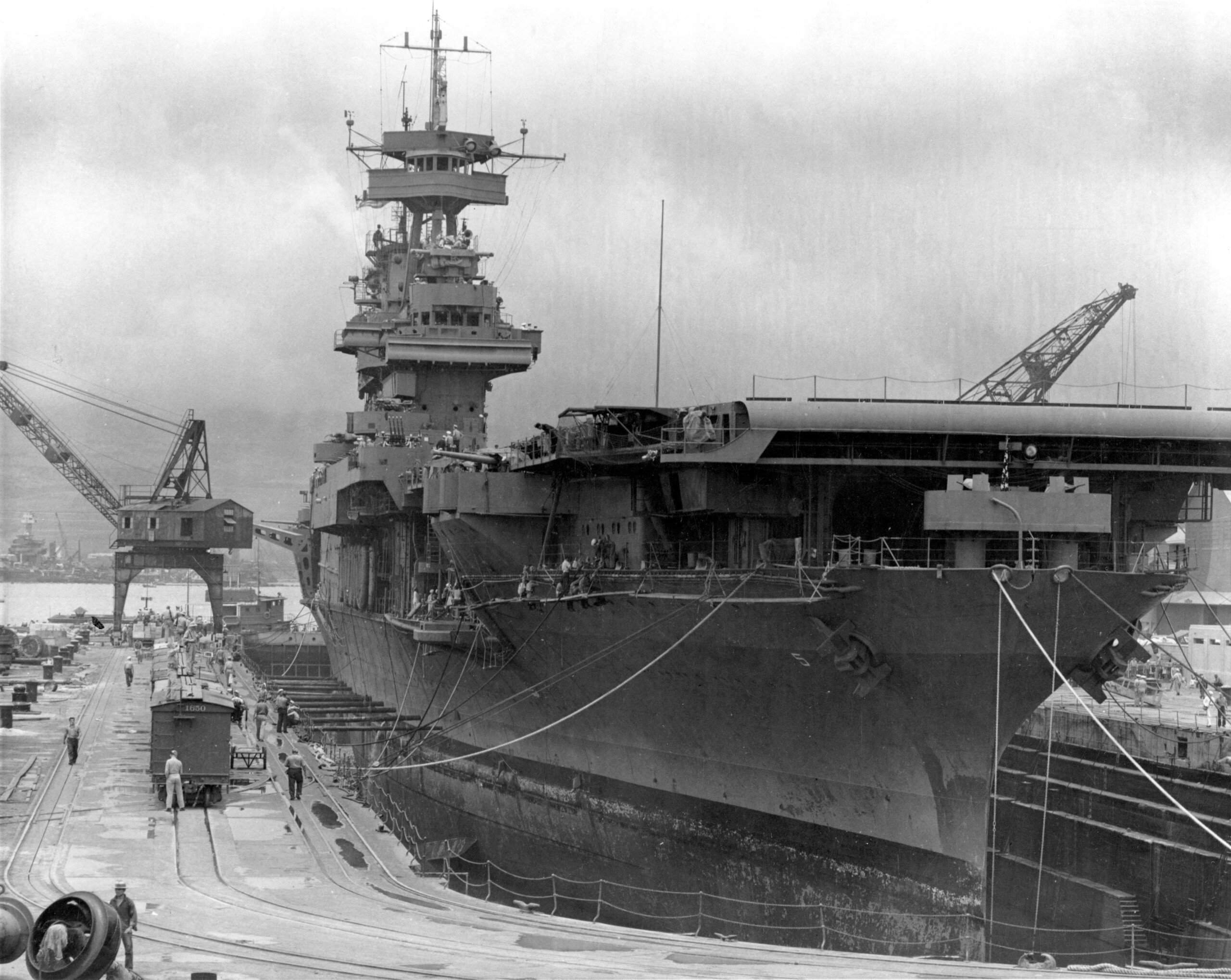 A large aircraft carrier is docked in a shipyard, surrounded by cranes and workers. The ship's hull shows signs of wear, and scaffolding is visible on the deck. The scene appears industrial and overcast.