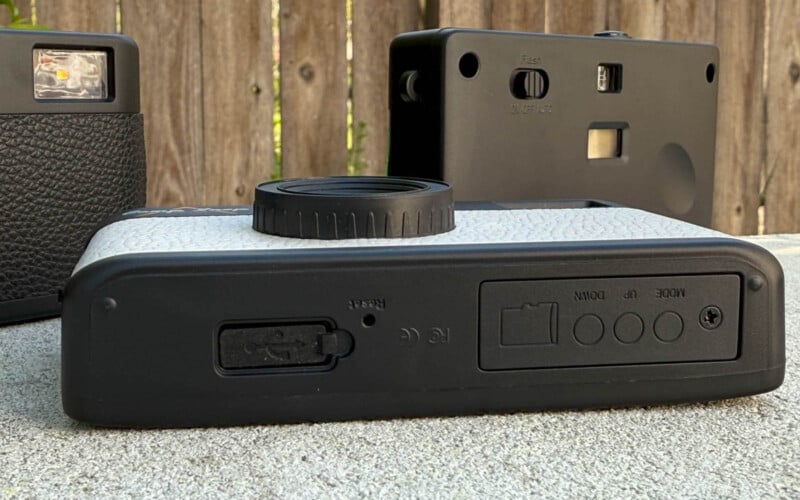 Close-up of three vintage cameras on a surface. The focus is on the bottom of a black and white camera with visible lens and controls. Two other cameras, one upright and one partially visible, are in the background against a wooden fence.