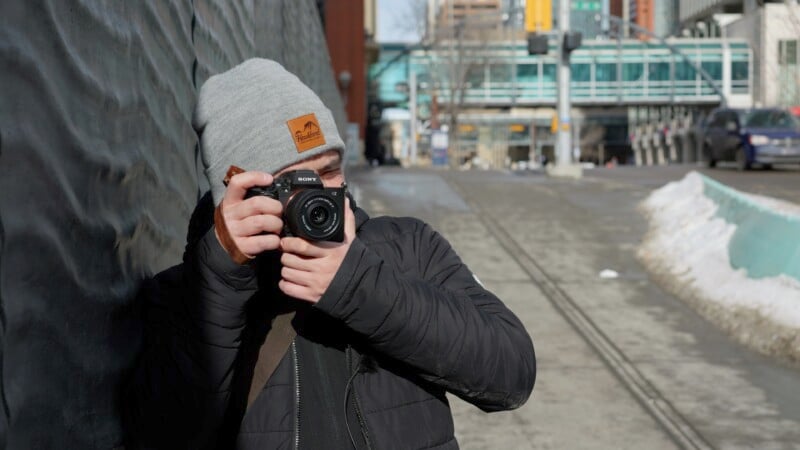 A person wearing a gray beanie and black jacket is holding a camera up to their face, taking a photo. They are standing on a city street with buildings and a few cars in the background. The street has patches of melting snow.
