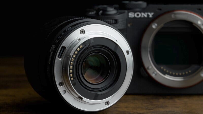Close-up of a camera lens facing a Sony camera body with no lens attached, placed on a dark wooden surface. The camera and lens are partially illuminated, highlighting the metallic mount and electronic contacts.
