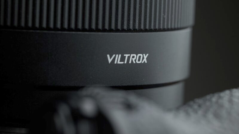 Close-up of a Viltrox camera lens, showing the brand name prominently on the side. The lens has a textured focus ring and a matte black finish, with part of a camera body visible in the foreground.