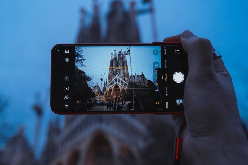 A hand holding a smartphone takes a picture of a large, intricate cathedral in the background. The phone screen displays the cathedral in focus, while the actual structure appears blurred. The sky is overcast with a bluish hue.